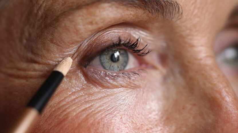 Close-up of a mature woman’s eye with a makeup pencil applying product to the outer corner, showing fine lines and natural lashes
