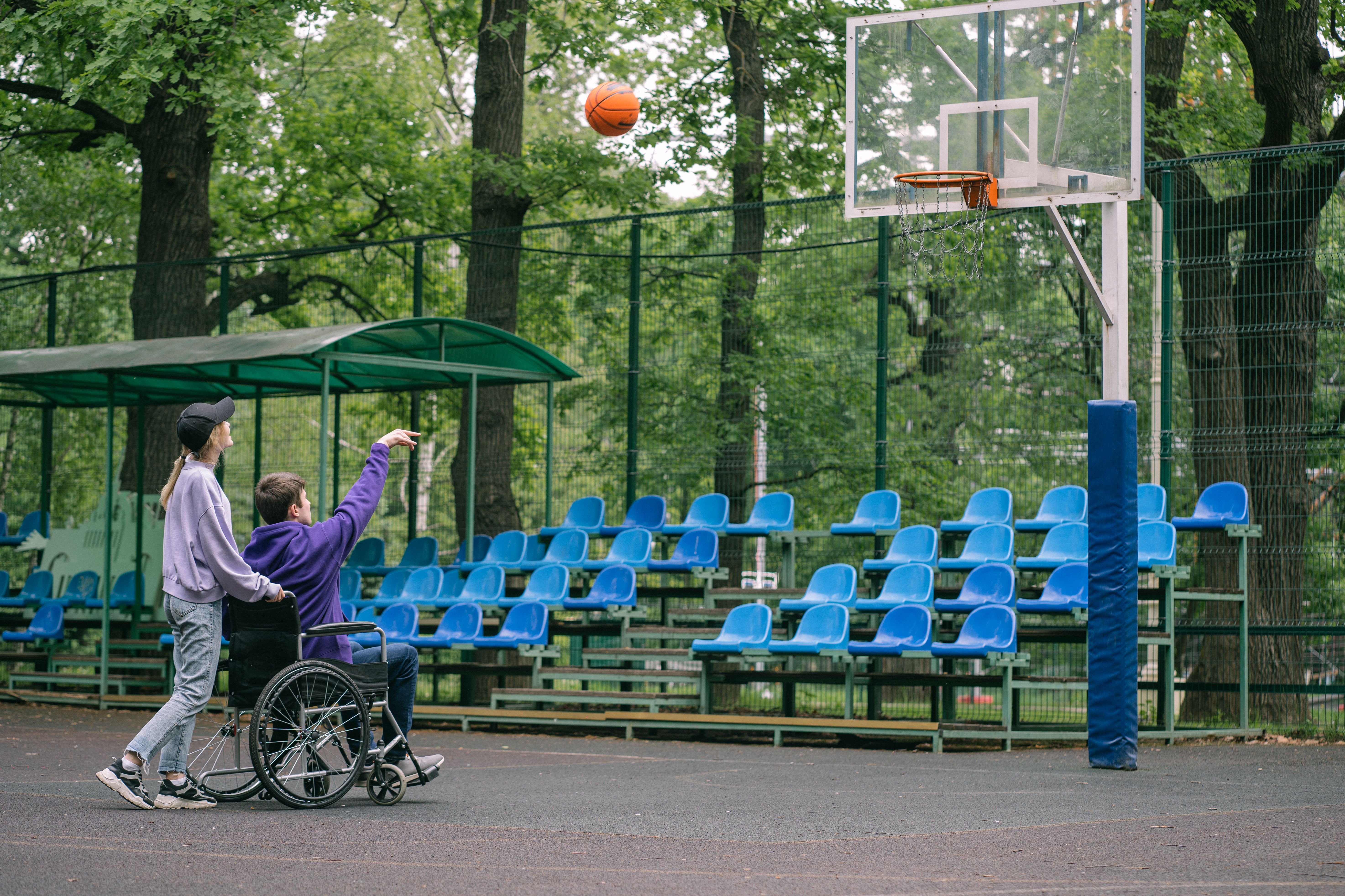 A person in a wheelchair playing basketball with a friend outdoors on an accessible court